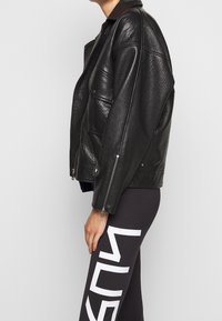 Svart läderjacka med en lätt strukturerad yta, med krage, dragkedjefickor och en avslappnad passform. Matchas med svarta leggings med vit text.