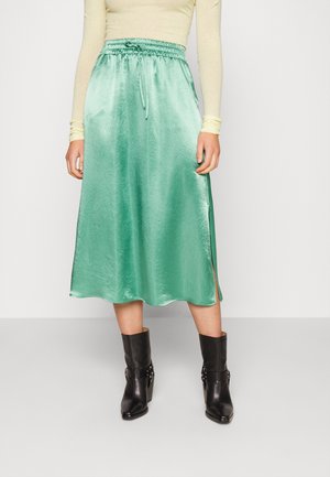 Femme portant un haut à manches longues jaune clair, une jupe midi vert menthe brillante avec taille à cordon, et des bottes noires à talons mi-mollet.