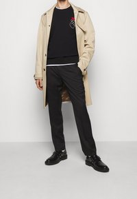 Un abrigo trench beige sobre una sudadera negra con un gráfico de mano y corazón, combinado con pantalones negros y zapatos negros. Texturas suaves en general.