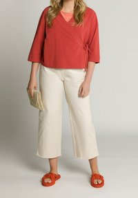 Rotes Wickeloberteil mit dreiviertel Ärmeln, cremefarbene Hosen mit weitem Bein und orangefarbene geknotete Sandalen. Das Modell hält eine gewebte Clutch-Tasche.