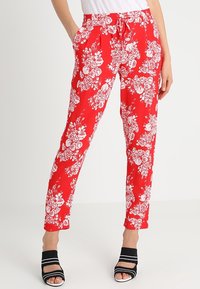 Rode drawstring broek met wit bloemenpatroon, gedragen door een staand persoon met handen in de zakken, gecombineerd met zwarte open teen schoenen.