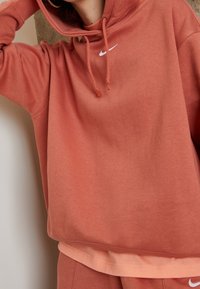 Rostfärgad hoodie i mjukt tyg, med en framficka, justerbara dragsnören och en liten vit Nike-logotyp på bröstet.
