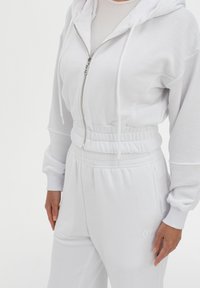 Felpa bianca con zip, cappuccio regolabile e vita elasticizzata, abbinata a pantaloni jogger bianchi con dettagli ricamati sul lato.
