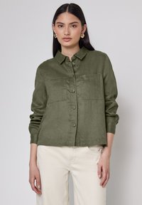 Camicia verde oliva con bottoni, colletto, due tasche sul petto e maniche lunghe, realizzata in un tessuto testurizzato, abbinata a pantaloni chiari.