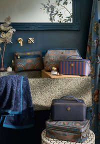 Patterned cosmetic bags in blue, orange, and cream; textured navy towel; stone countertop; ornate mirror; floral curtain.