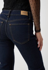 Vaqueros de mezclilla de color azul oscuro con un corte slim, que presentan costuras doradas, un parche de cuero texturizado con el logo y dos bolsillos tipo parche en la parte posterior.