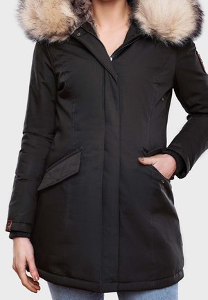Femme portant un manteau d'hiver noir avec une capuche bordée de fourrure beige, les mains dans les poches, et une fermeture éclair à l'avant.
