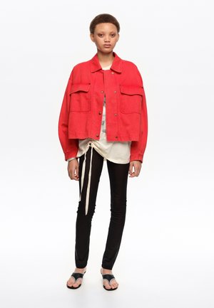 Młoda osoba w luźnej czerwonej kurtce, kremowym topie z wiązaniami, czarnych dopasowanych spodniach i czarnych sandałach z odkrytymi palcami, stojąca na białym tle.