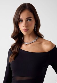 Klares Kristallhalskette mit einer Mischung aus rechteckigen und abgerundeten Steinen; kombiniert mit passenden Ohrringen. Modell trägt ein schwarzes, schulterfreies Oberteil.