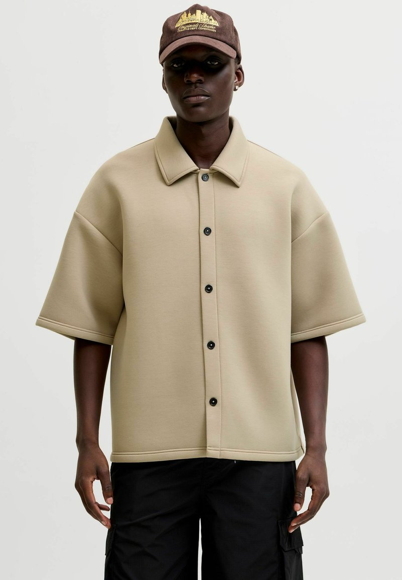 Personne à la peau foncée portant une chemise beige à manches courtes boutonnée, un pantalon cargo noir et une casquette marron avec un texte jaune et un motif de skyline.