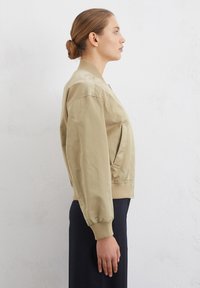 Blouson beige avec des poignets et un ourlet côtelés, fermeture éclair à l'avant, poches latérales et texture lisse et brillante. Modèle vu de côté.