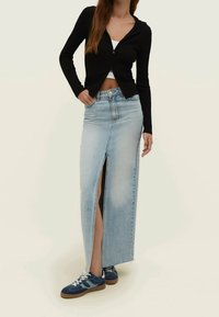 Kobieta ubrana w czarną bluzę na zamek, białą crop top, jasnoniebieską maxi spódnicę dżinsową z przednim rozcięciem i niebieskie sneakersy z białymi sznurowadłami.