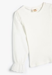 Chemise blanche à manches longues côtelée, avec un col rond et un tissu extensible. L'extrémité des manches présente un design en forme de scallop avec des accents de trous.