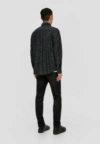 Zwarte, gestructureerde, langemouwen shirt met een kraag, weergegeven van achteren. Gecombineerd met zwarte broek en platte zwarte schoenen.