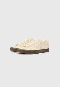 Beige canvas sneakers with a rubber sole, rounded toe, and flat laces. Features a textured upper and subtle logo detail on the side.