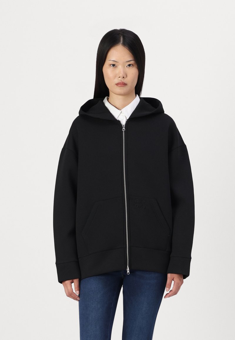 Sweat à capuche noir zippé en tissu doux, avec une grande capuche, des poches avant et un logo en relief minimaliste sur la poitrine.