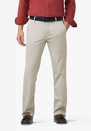 Uomo che indossa pantaloni beige aderenti, cintura intrecciata nera, mocassini neri e una camicia rossa a maniche lunghe con maniche arrotolate.