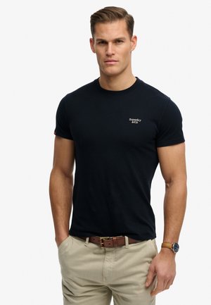 Hombre con una camiseta negra ajustada de Superdry & Co, pantalones beige con cinturón marrón y un reloj de pulsera, de pie frente a un fondo liso.