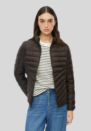 Junge Frau trägt eine schwarze gesteppte Jacke über einem weiß-schwarz gestreiften Hemd und blauen Jeans, steht vor einem grauen Hintergrund.
