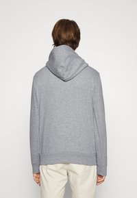 Person med kort brunt hår som bär en enkel grå huvtröja och ljusbeige byxor, sedd bakifrån mot en enhetlig bakgrund.