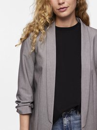 Vrouw met krullend blond haar, met grote oorbellen, een grijze blazer met opgerolde mouwen, een zwart shirt en een blauwe spijkerbroek, tegen een witte achtergrond.
