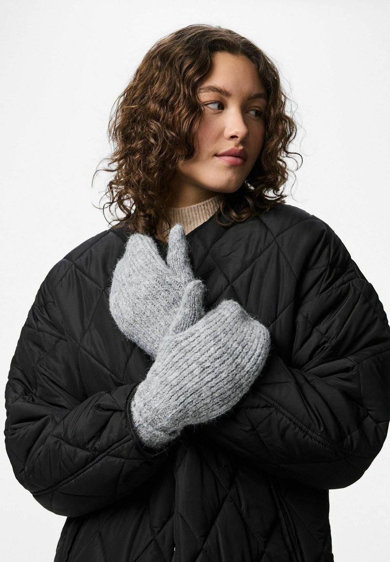 Grå strikkede vanter med en blød, tekstureret afslutning, parret med en sort quiltet jakke. Vanterne er oversized og hyggelige.