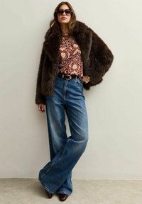 Donna con un'oversize giacca di pelliccia marrone scuro, camicetta a fantasia, pantaloni jeans blu a gamba larga, cintura scura, scarpe marroni a punta e occhiali da sole, in piedi con sicurezza.