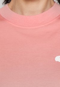 T-shirt en coton rose avec un col rond, une texture lisse et un détail graphique blanc sur le côté. Design simple et uni sans motifs.