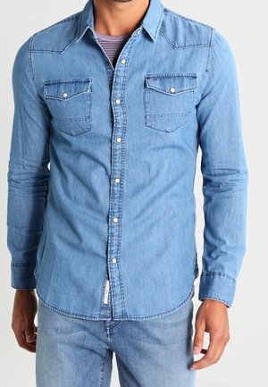 Homme portant une chemise en denim bleu clair à boutons sur un T-shirt violet rayé sans col et un jean bleu clair assorti.