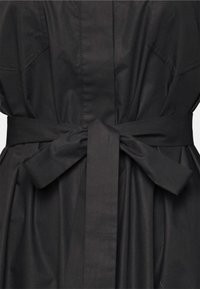 Musta mekko, jossa on solmittu vyötärönauha, sileä pinta ja rento siluetti. Saumojen kohdalla näkyy yksityiskohtaiset tikkausompeleet.