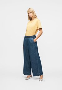 Donna che indossa una maglietta gialla a maniche corte infilata in pantaloni di jeans blu a gamba larga e tacchi bianchi, in piedi con una mano in tasca.