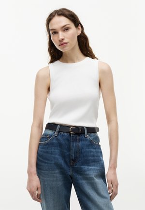 Γυναίκα με μακριά καστανά μαλλιά φοράει λευκό αμάνικο τοπ, μπαίνει μέσα σε ψηλόμεσο μπλε τζιν με μαύρη ζώνη, στέκεται μπροστά σε απλό φόντο.