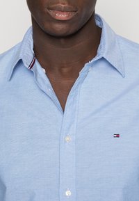 Hombre vestido con una camisa de cuello azul claro, con un pequeño logo rojo, blanco y azul en el pecho, parcialmente abotonada y con un detalle de cuello interior a rayas.