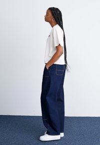 T-shirt branca de manga curta com logo bordado, combinado com jeans escuros de corte largo e ténis brancos. Cabelo longo em tranças.