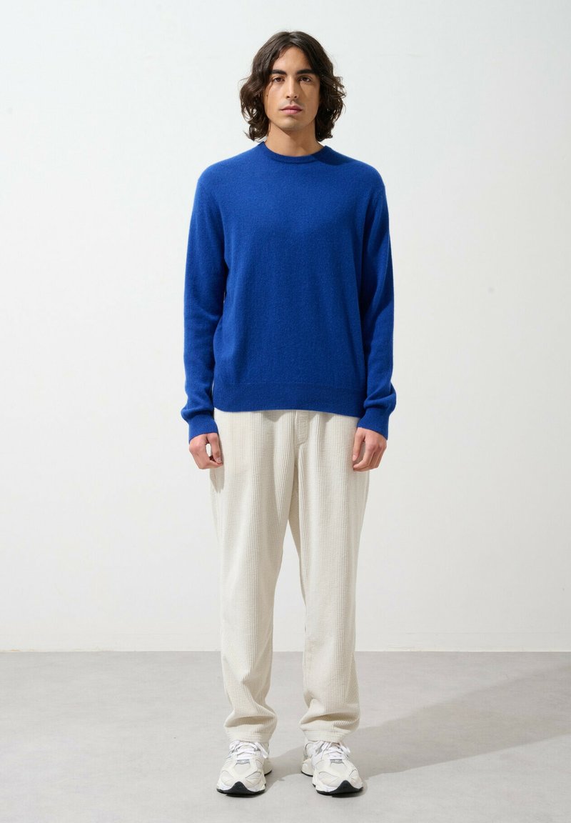 Maglione blu in lana con scollatura rotonda, maniche lunghe e polsini a coste, abbinato a pantaloni cremosi con texture e sneakers bianche.