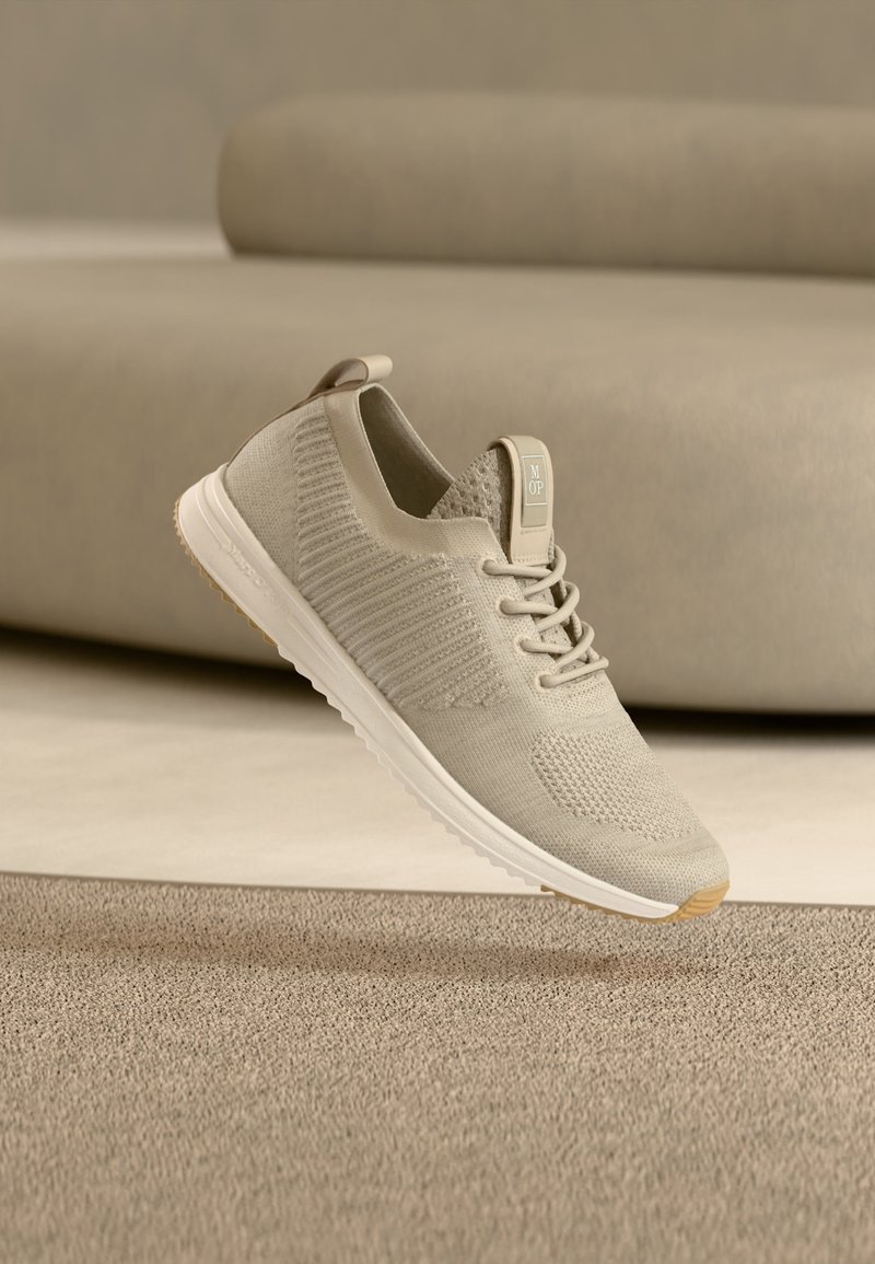 Baskets tricotées beige clair avec semelle blanche, lacets et languette, montrées flottant au-dessus d'une surface texturée avec un canapé flou en arrière-plan.