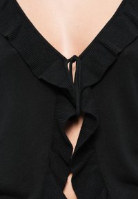 Μαύρη πλεκτή ζακέτα με λεπτομέρειες βολάν και κλείσιμο με δέσιμο, μισάνοιχτη για να φαίνεται το δέρμα από κάτω.