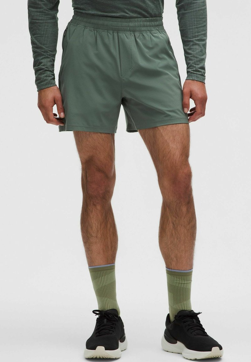 Mann in grünen Sportshorts, passenden grünen Socken, schwarzen Laufschuhen und einem langärmligen, strukturierten grünen Oberteil, steht vor einem einfarbigen Hintergrund.