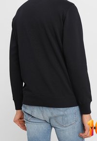 Felpa nera con collo rotondo e polsini a costine, abbinata a jeans azzurri. Il tessuto sembra morbido con una texture liscia.