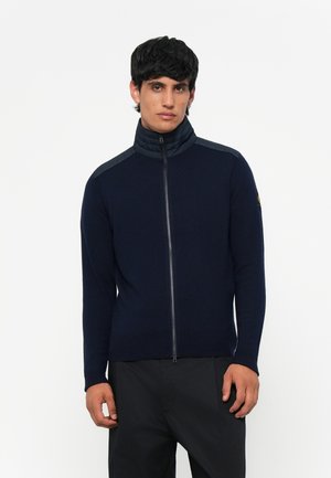 Navy zip-up sweater with a high collar, featuring a smooth texture, knit body, and contrasting quilted shoulder accents.