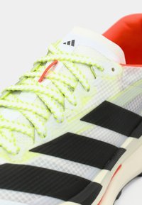 Αθλητικό παπούτσι σε λευκό και μαύρο με επάνω μέρος από δίχτυ, φωσφορίζοντα κίτρινα κορδόνια και κόκκινες λεπτομέρειες. Διαθέτει τρεις μαύρες ρίγες στο πλάι.