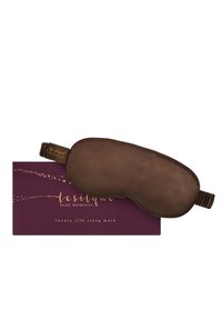 Maschera da sonno di lusso in seta marrone poggiata su una scatola viola scuro con scritte dorate e design a stella.