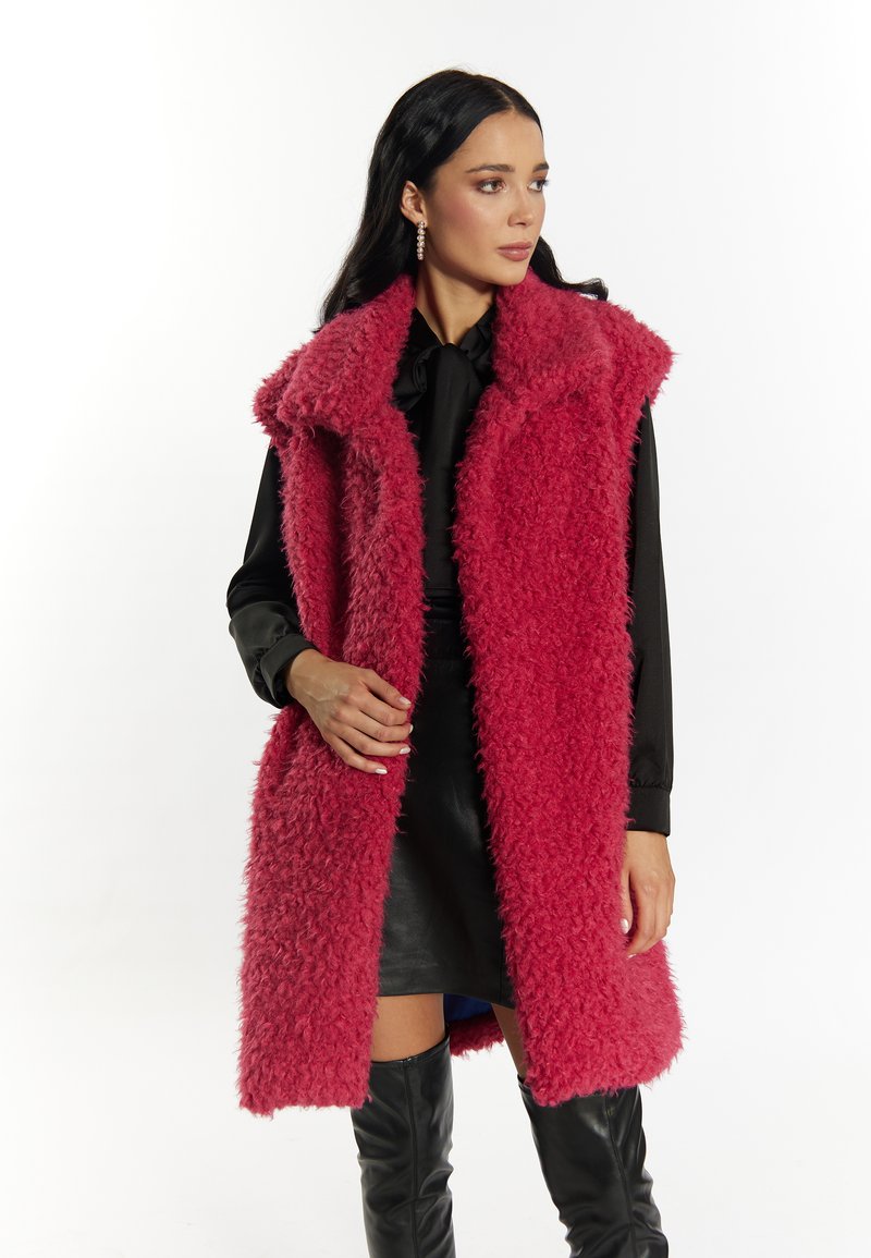 Lang mouwloos jasje van gestructureerd roze nepbont, met een brede kraag en een open voorkant. Gecombineerd met een zwarte blouse en een leren rok.