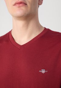 Burgunderfarget v-hals t-skjorte laget av myk bomull, med en liten hvit brodert logo på brystet. Kjempefin tekstur med avslappet passform.
