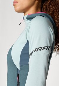 Hele sinise ja tumeteal värvi sportlik jakk, millel on lukuga kapuuts, tekstuuriga kangas ja must "DYNAFIT" logo varrukal. Ka roosad aktsendid.