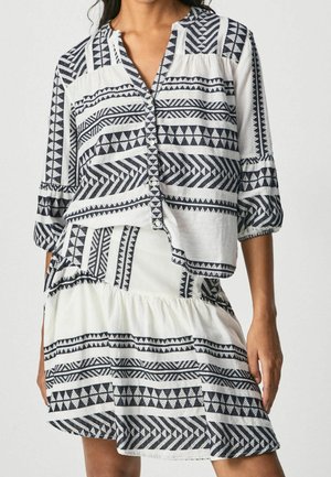 Frau trägt eine weiße Bluse und einen Rock mit schwarzen geometrischen Tribal-Mustern, Dreiviertelärmel und durchgeknöpftem Vorderteil.