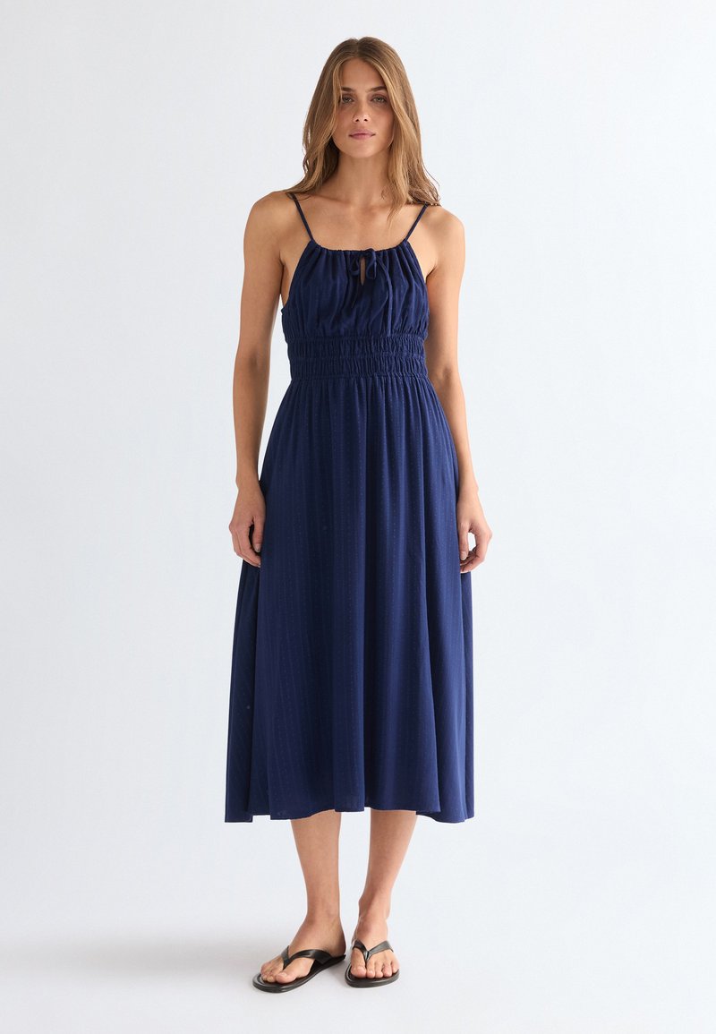 Γυναίκα που στέκεται φορώντας ένα μπλε σκούρο midi φόρεμα χωρίς μανίκια με σφηκοφωλιά στη μέση και μαύρες σαγιονάρες σε λευκό απλό φόντο.