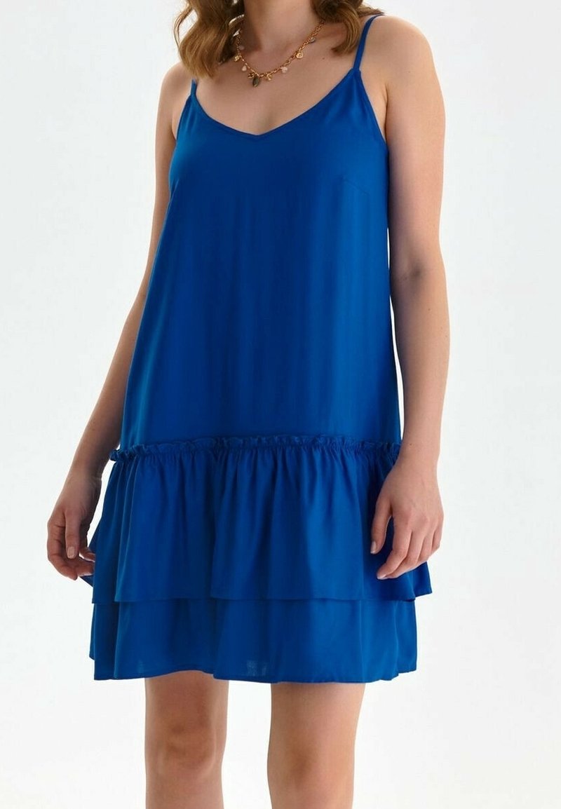 Mujer vestida con un vestido sin mangas de color azul royal, con tirantes finos y un dobladillo de volantes en capas, complementado con un collar de oro.