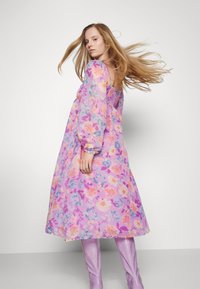 Vestido midi floral em tom pastel de roxo, com mangas longas, corpete franzido e padrões vibrantes de flores coloridas. Usado com botas rosa brilhantes.