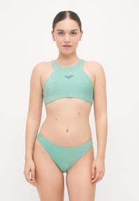 Világoszöld bikini szett csillogó textúrával. A felsőrész magas nyakú és logóval rendelkezik, míg az alsó része alacsony derékú kialakítású.